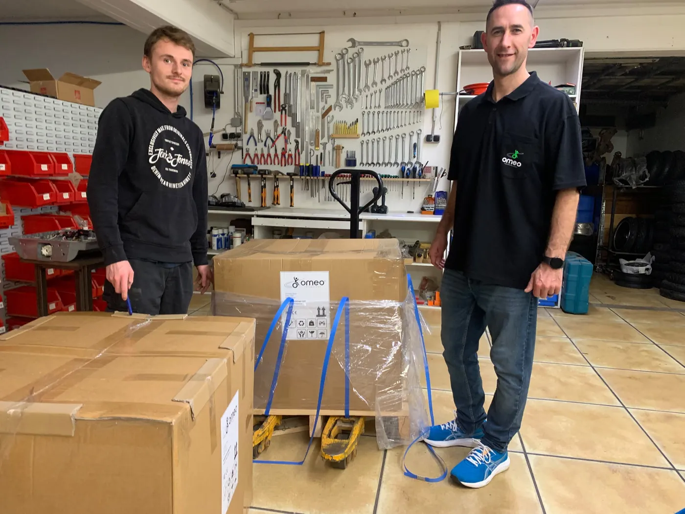 Zwei PT-Pro-Mitarbeiter neben originalverpackten Omeo-Versandkisten von Omeo Technology in der Werkstatt